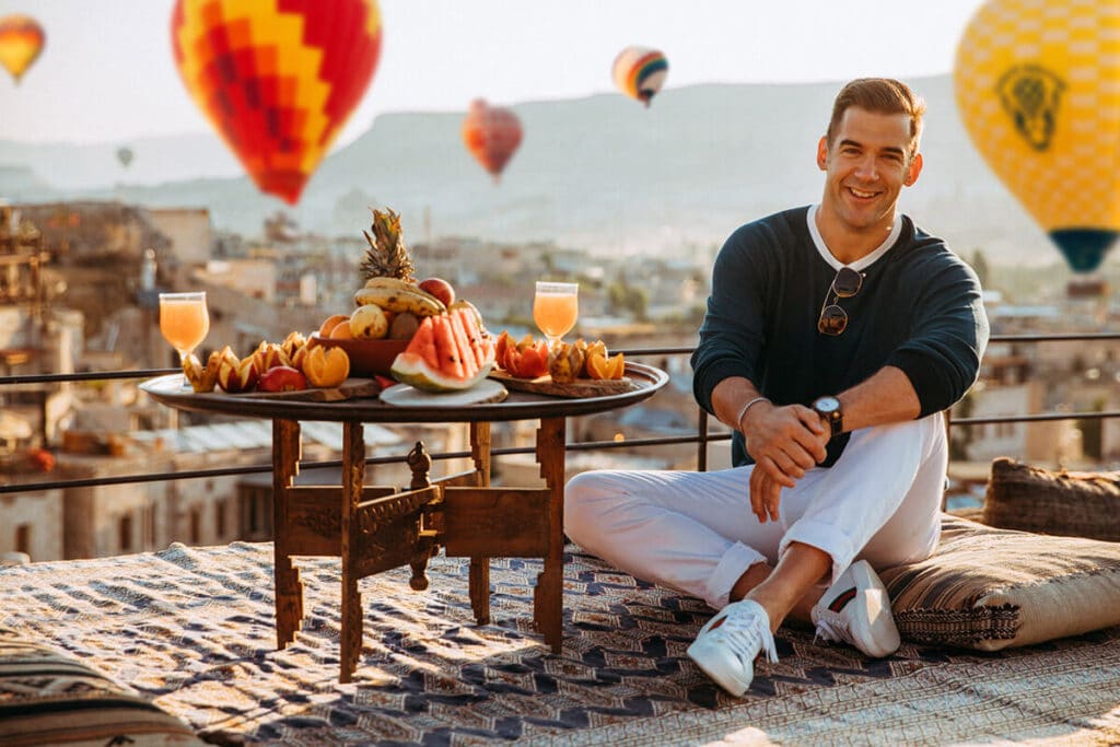 Lewis Howes sitting beside a vibrant rooftop breakfast spread in Cappadocia with hot air balloons rising behind him — reflecting warmth, grounded presence, and lifestyle storytelling through personal brand photography