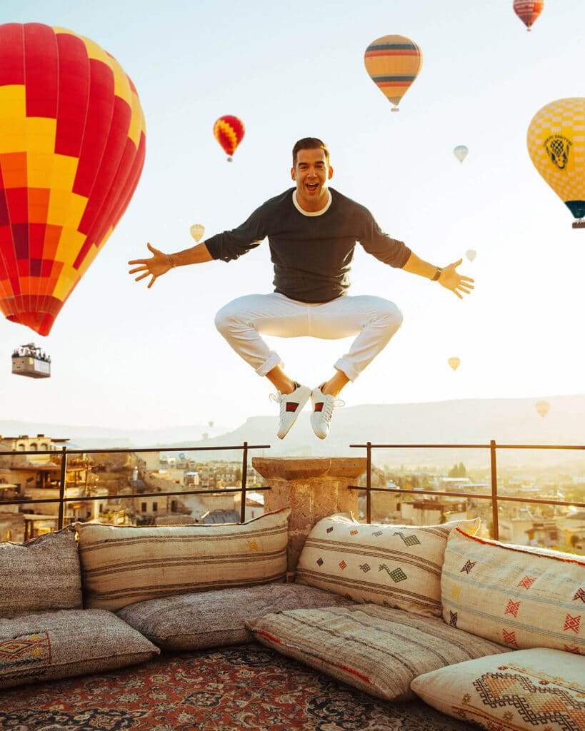 Lewis Howes jumping with joy on a rooftop in Cappadocia with hot air balloons in the background — expressing freedom, global presence, and personal brand expansion through Elevated Realism™