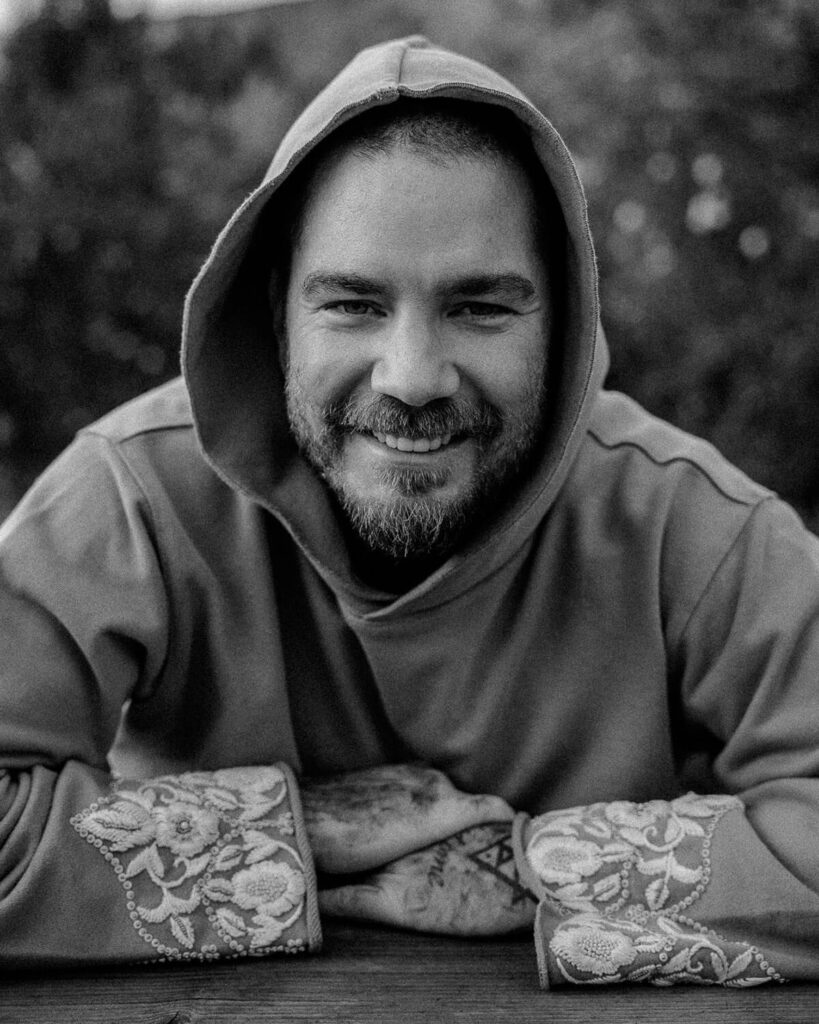 Black-and-white portrait of tattoo artist Balazs laughing outdoors on a wooden bench, with mountain views in the background