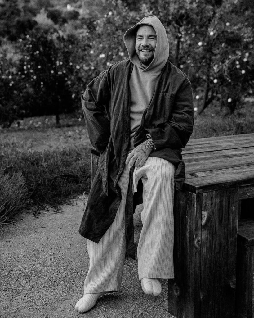 Candid black-and-white portrait of Balazs, visionary tattoo artist, in natural light wearing a hooded sweatshirt and expressing warmth