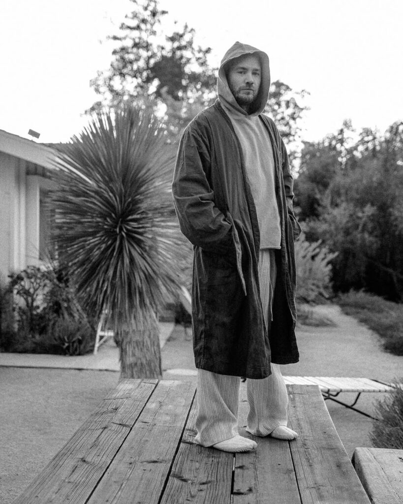 Full-body portrait of tattoo artist Balazs standing in layered robes on a deck, embodying mystical presence and grounded creative flow