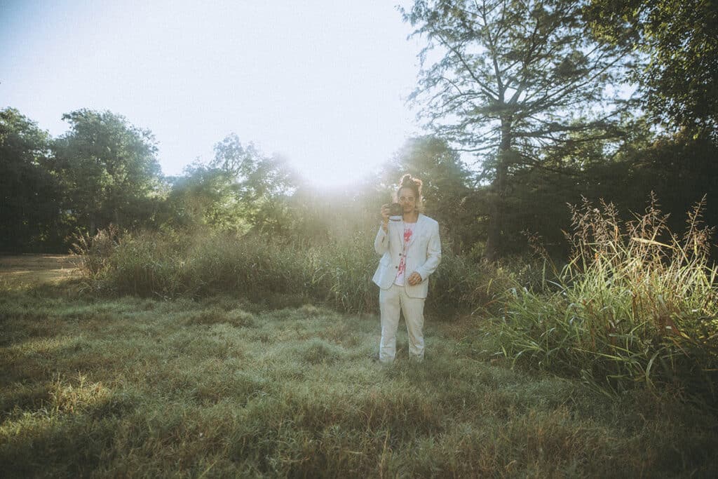 Nick Onken standing in a dew-covered field at sunrise, holding his camera in a white suit, embodying creative clarity and presence on the path